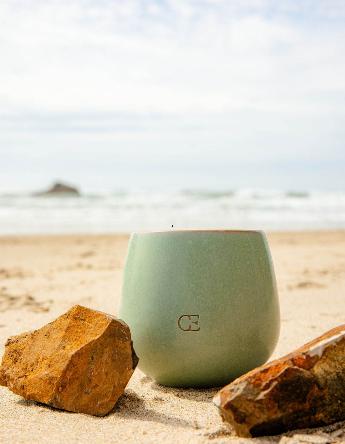 A Candle UNWIND by Cozy Earth sits on a sandy beach surrounded by rocks, with the ocean and cloudy sky in the background.