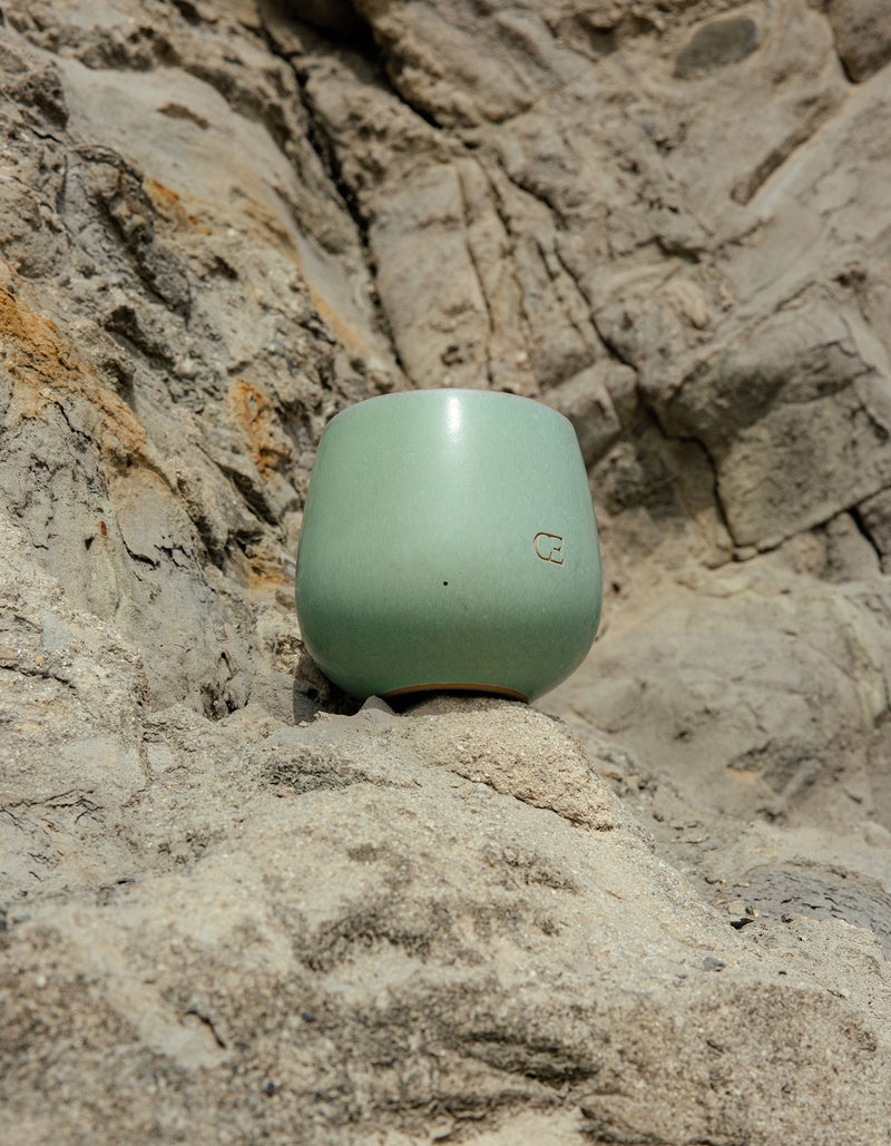 The Candle UNWIND by Cozy Earth, a light green ceramic cup with a small logo, rests on textured rocks under natural sunlight.
