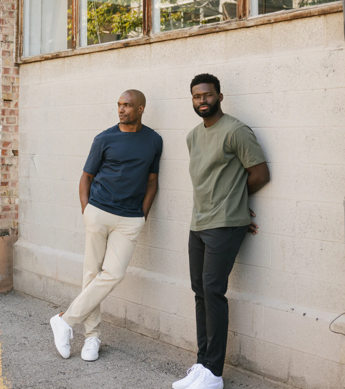 Two men stand against a brick wall wearing Cozy Earth Add-Day Tees and Everywhere Pants.