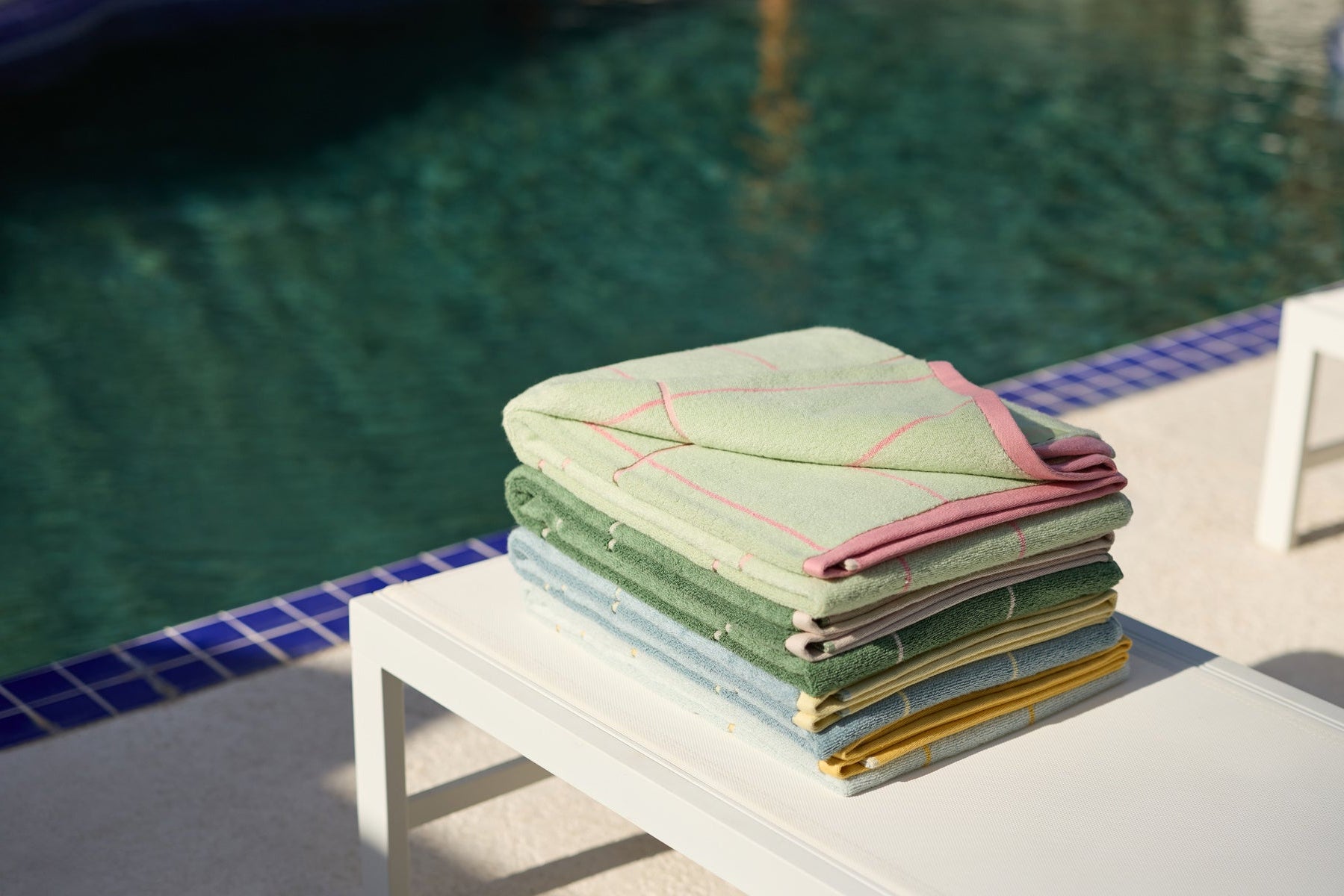 A neatly folded stack of colorful towels sits on a white table beside a swimming pool with blue tile edging, bathed in sunlight.