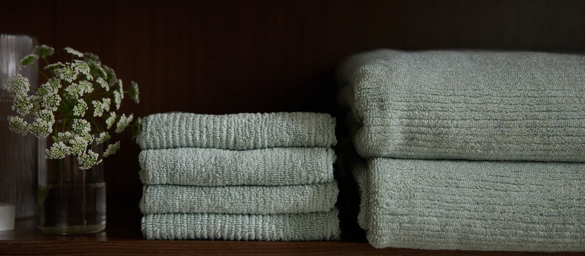 A hand places a folded gray Cozy Earth Complete Ribbed Terry Bath Bundle towel atop a neatly stacked pyramid of towels, set before a marble-tiled wall.|Color:Charcoal