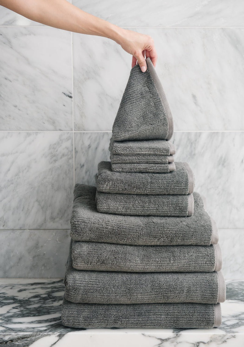 A hand places a folded gray Cozy Earth Complete Ribbed Terry Bath Bundle towel atop a neatly stacked pyramid of towels, set before a marble-tiled wall.