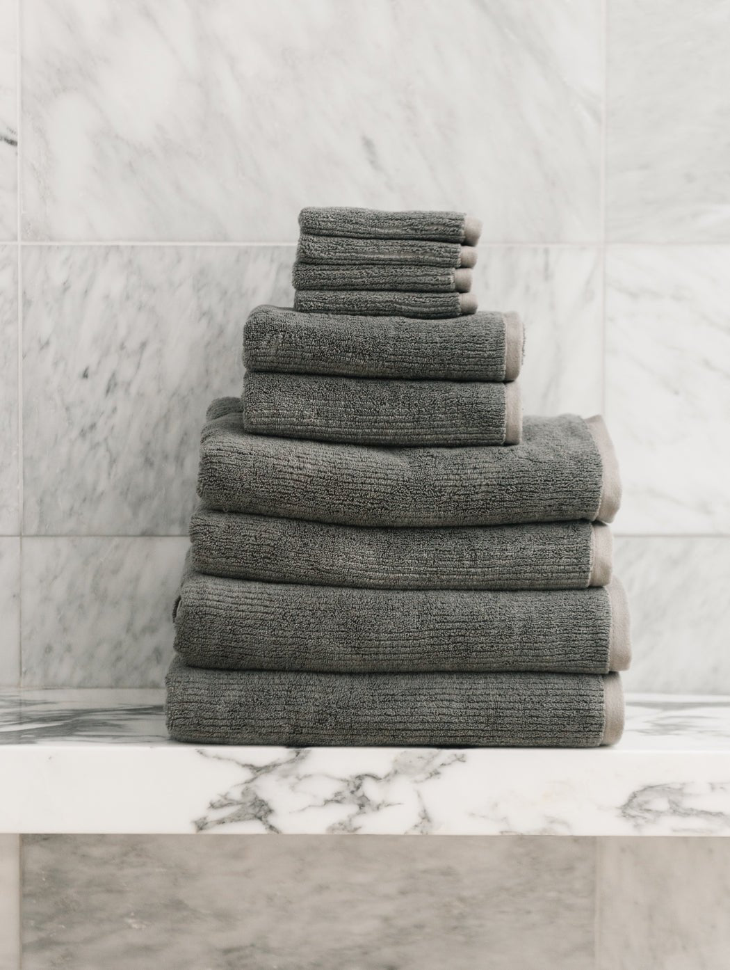 The Cozy Earth Complete Ribbed Terry Bath Bundle, featuring gray towels in assorted sizes, is displayed neatly on a white marble shelf with a marble-tiled wall backdrop.