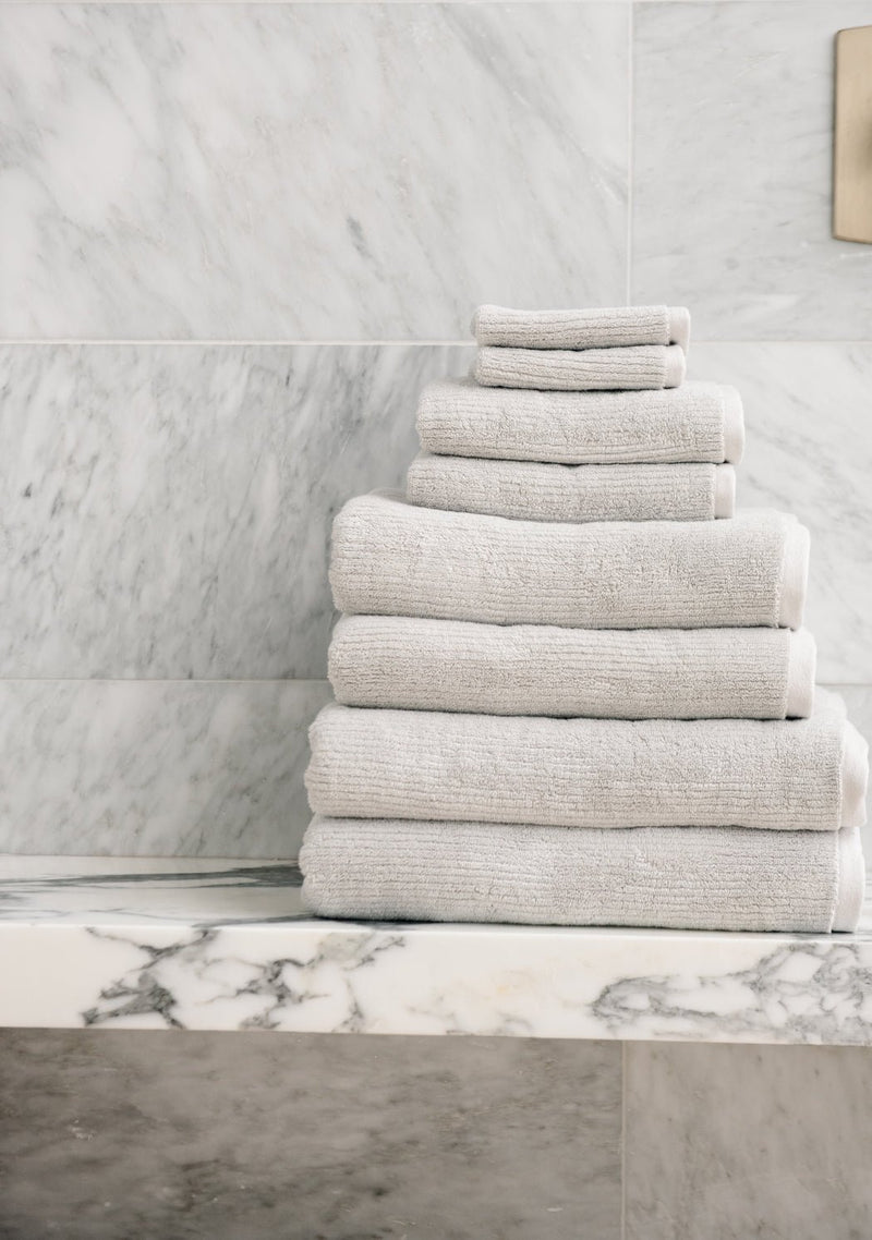 The Complete Ribbed Terry Bath Bundle by Cozy Earth is neatly stacked on a marble shelf in a bathroom with light gray marble walls.