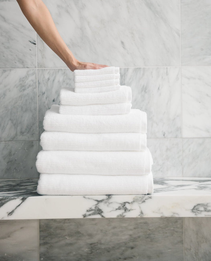 A hand places a neatly folded Cozy Earth Complete Ribbed Terry Bath Bundle towel atop a pyramid-shaped stack on a marble countertop, with a marble-tiled wall in the background.