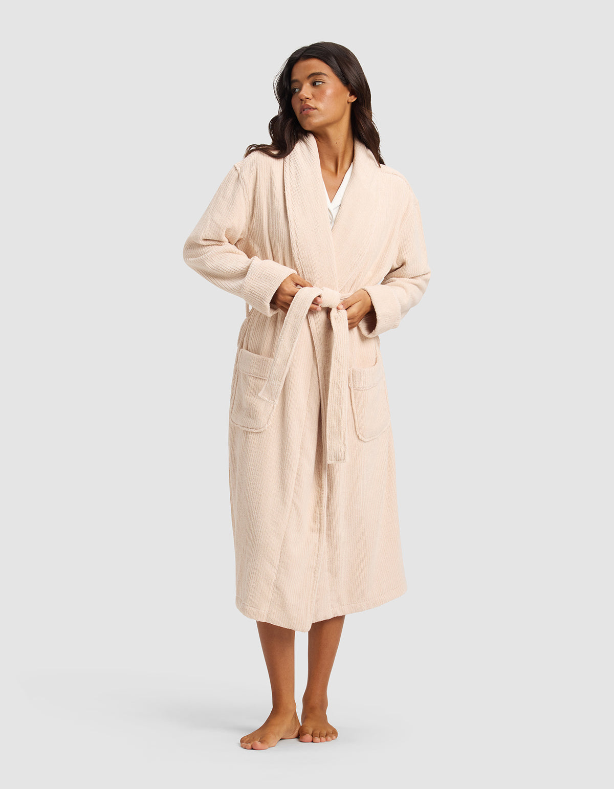 A woman stands barefoot against a plain light gray background, wearing Cozy Earth's Ribbed Terry Bath Robe in light beige featuring long sleeves, large front pockets, and a belt.