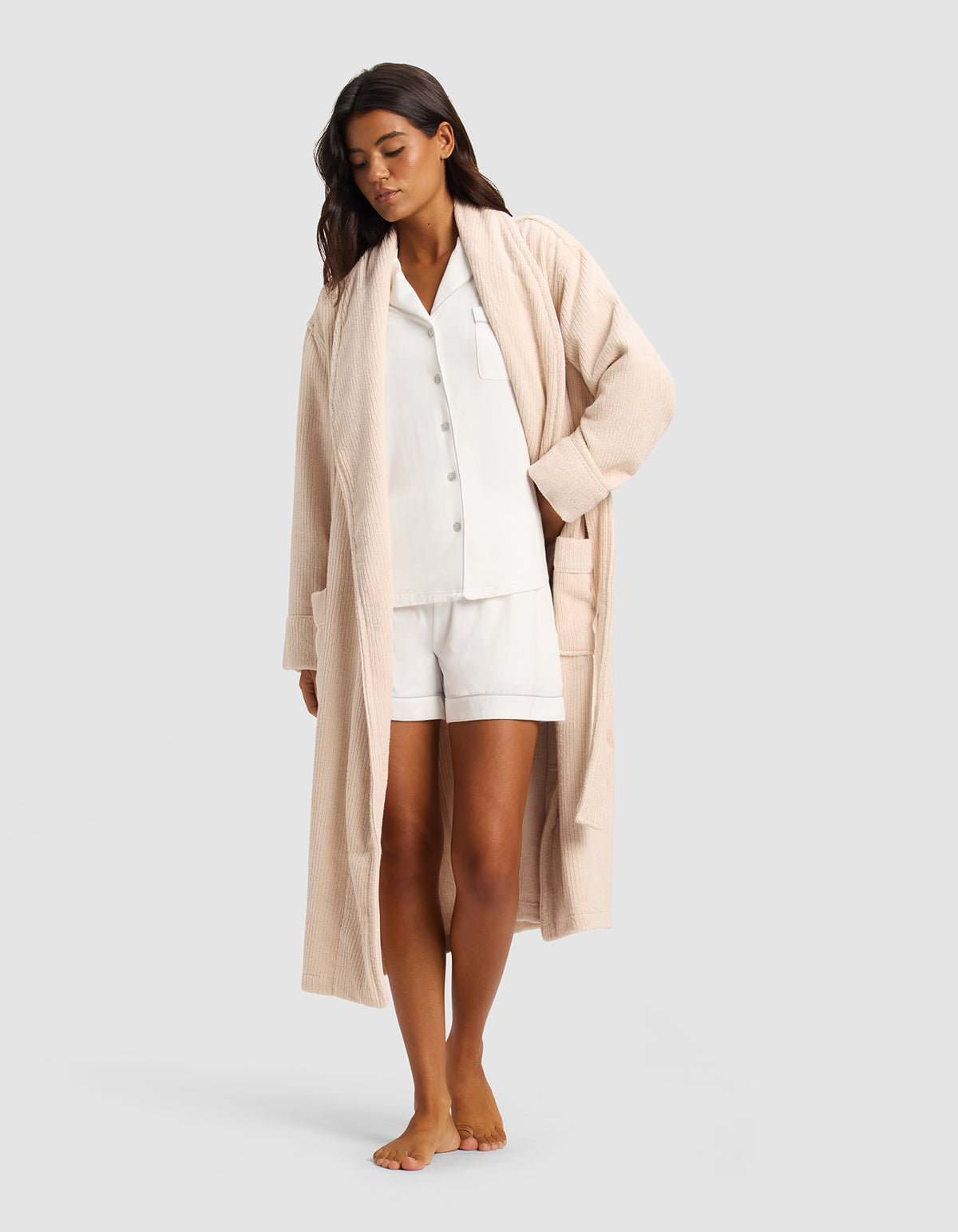 A woman stands barefoot in a white pajama set with shorts, layered under the Cozy Earth Ribbed Terry Bath Robe in light beige. She looks down with one hand in her robe pocket against a plain light gray background.