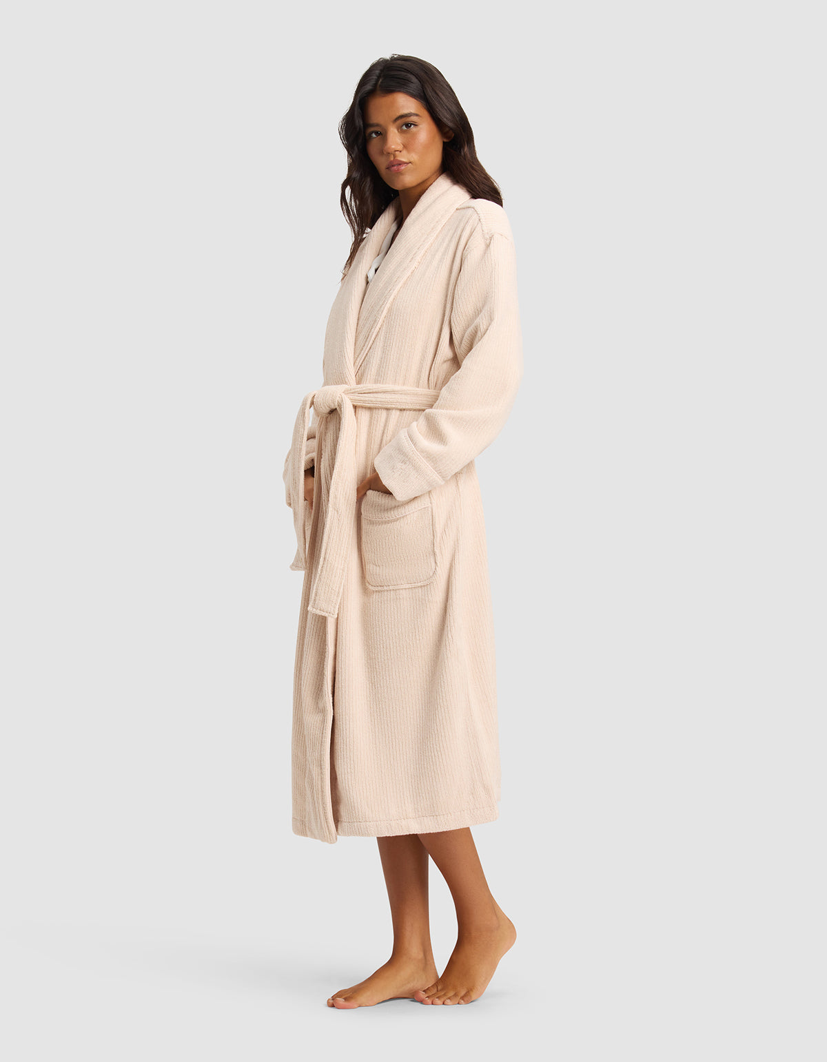 A woman stands barefoot in the Cozy Earth Ribbed Terry Bath Robe, a long light beige robe with a belt and two front pockets. She faces slightly to the side against a plain light gray background.