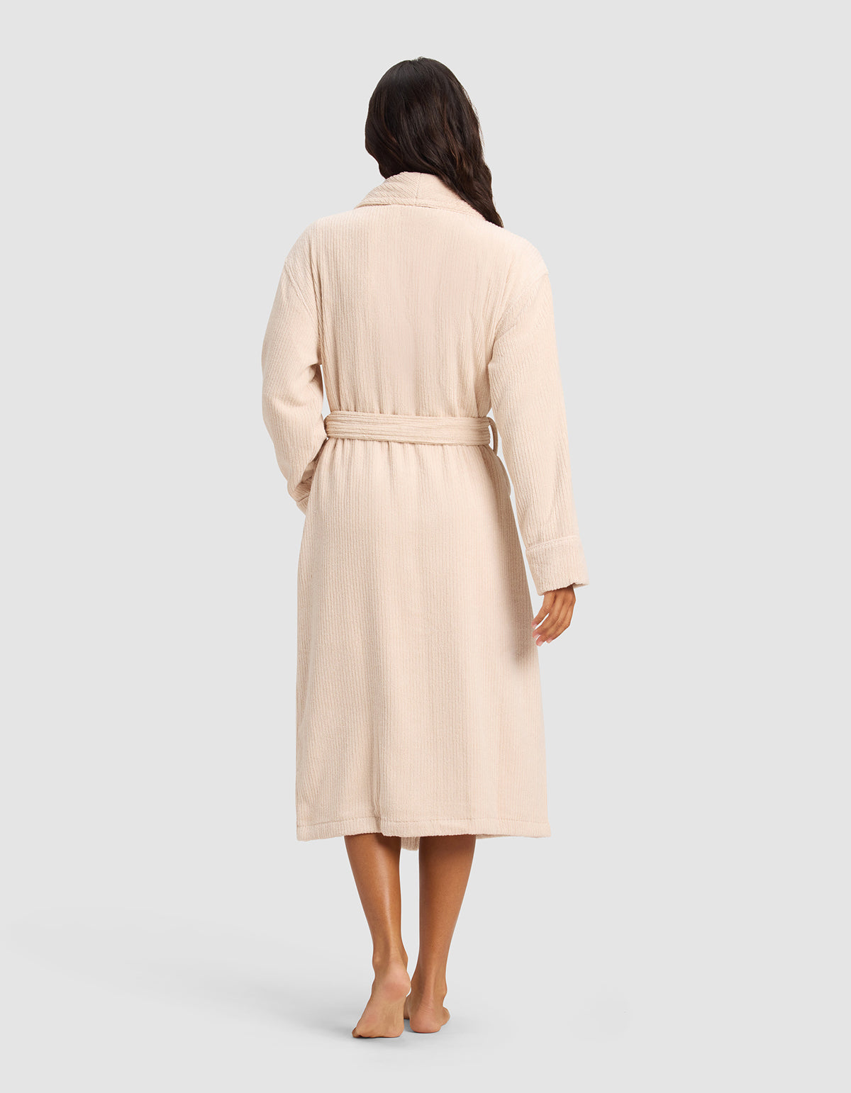 A woman with long dark hair stands barefoot, facing away, wearing the Cozy Earth Ribbed Terry Bath Robe in beige. The robe is long-sleeved and tied at the waist. The background is plain and light gray.