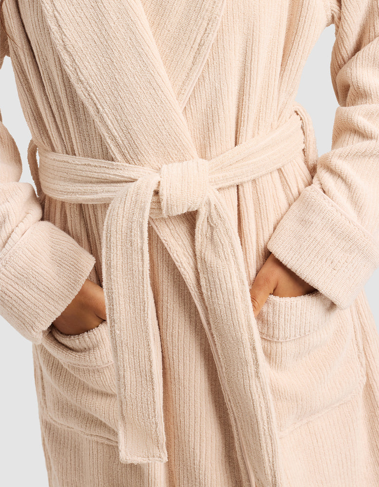 A person wears the Cozy Earth Ribbed Terry Bath Robe in beige, featuring a textured finish, waist belt, and hands in the front pockets.