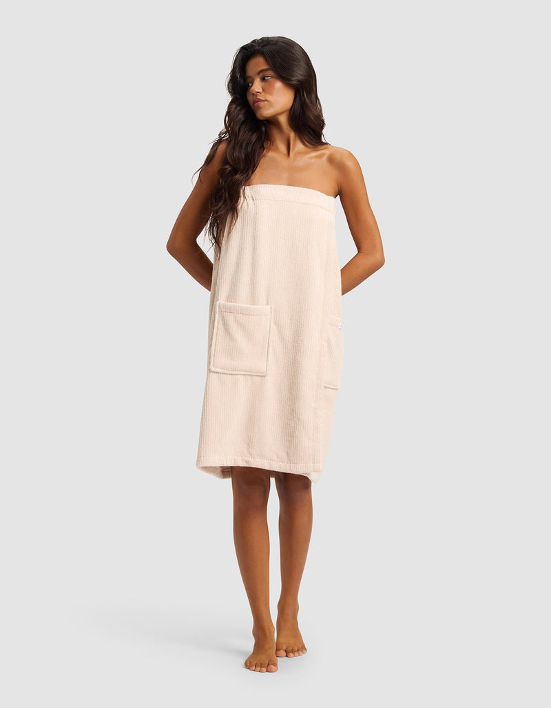 A woman with long dark hair stands barefoot against a plain light gray background, wearing the Cozy Earth Ribbed Terry Bath Wrap in light beige with two front pockets.