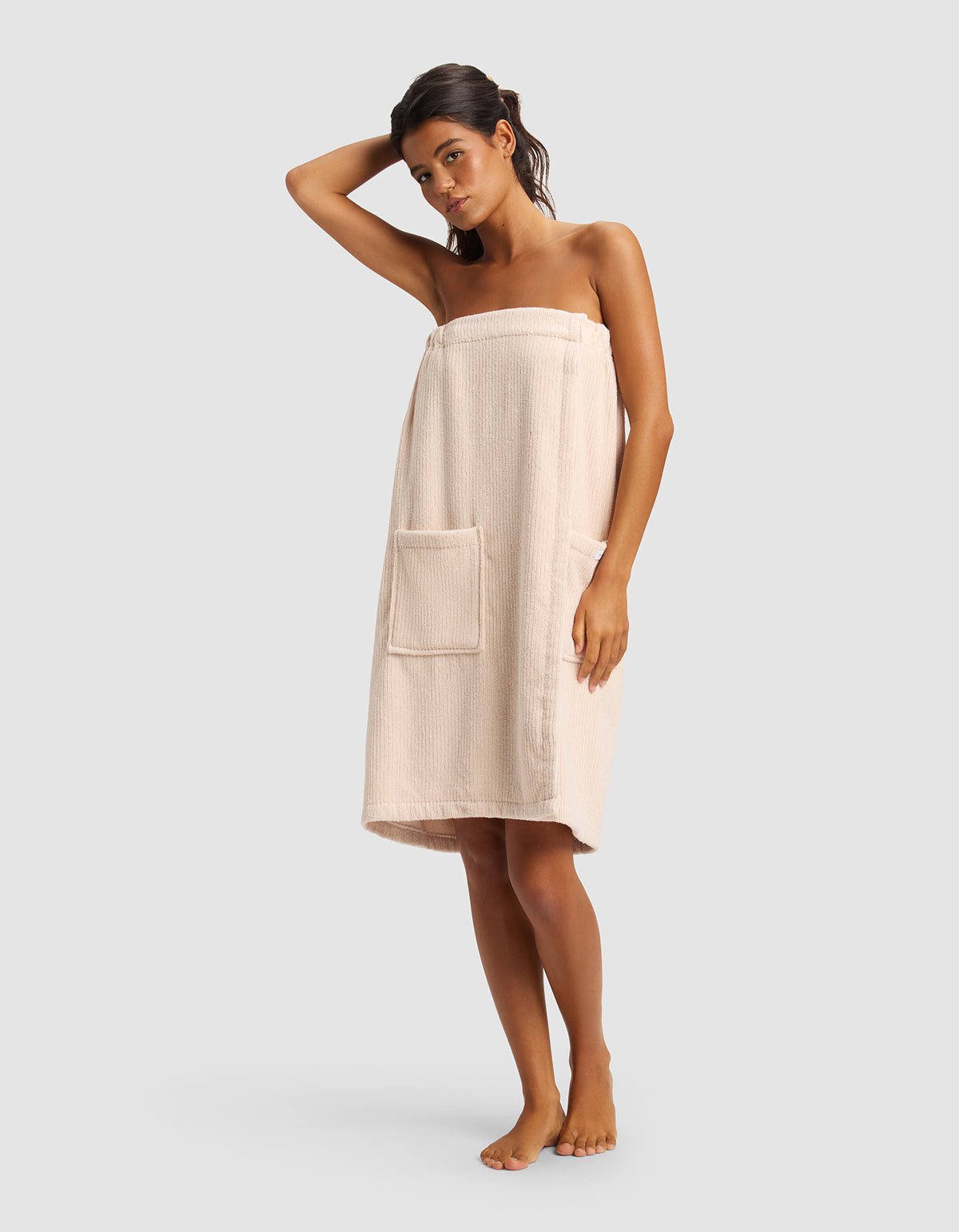 A woman stands barefoot against a plain background, wearing the Cozy Earth Ribbed Terry Bath Wrap with a large pocket, and has one arm raised to touch her head.