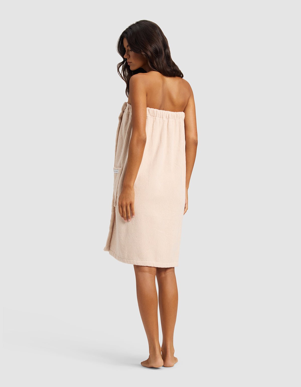 A woman with long dark hair, barefoot and facing slightly away, wears the Cozy Earth Ribbed Terry Bath Wrap in beige against a plain light gray background.