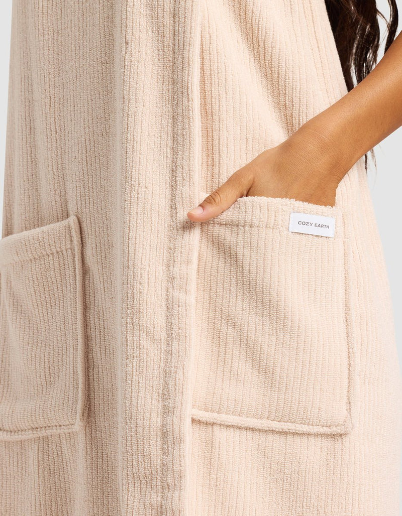 Someone wears Cozy Earth's Ribbed Terry Bath Wrap in beige, featuring ribbed fabric and large front pockets—one hand rests inside a pocket with the Cozy Earth label showing.