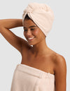 A smiling woman stands against a light gray background, wearing a beige towel wrap and the Cozy Earth Ribbed Terry Hair Towel in matching beige, with one arm raised and bent behind her head.|Color:Petal