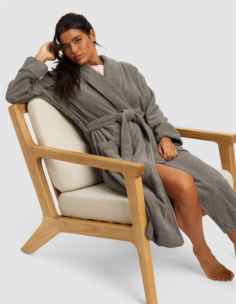 A woman with long dark hair sits barefoot on a modern wooden chair, wearing the Cozy Earth Ribbed Terry Bath Robe in gray. She rests her elbow on the armrest and gazes at the camera with a relaxed expression.