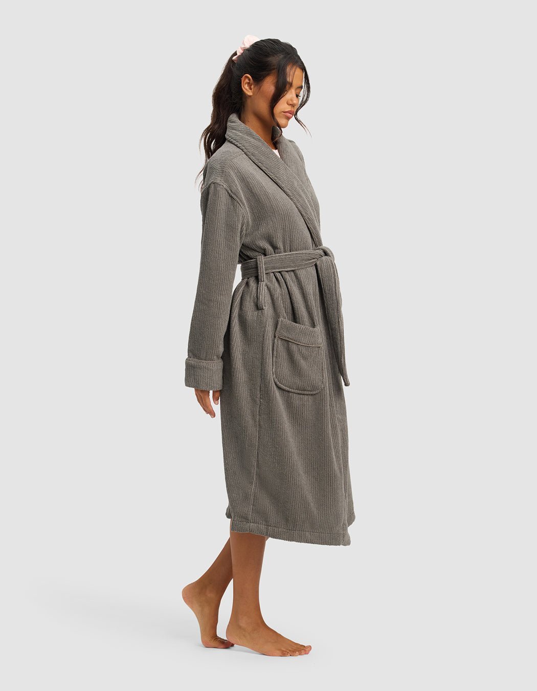 A woman with long dark hair stands barefoot in profile against a light background, calmly resting her hands in the pockets of a gray, knee-length Ribbed Terry Bath Robe by Cozy Earth.