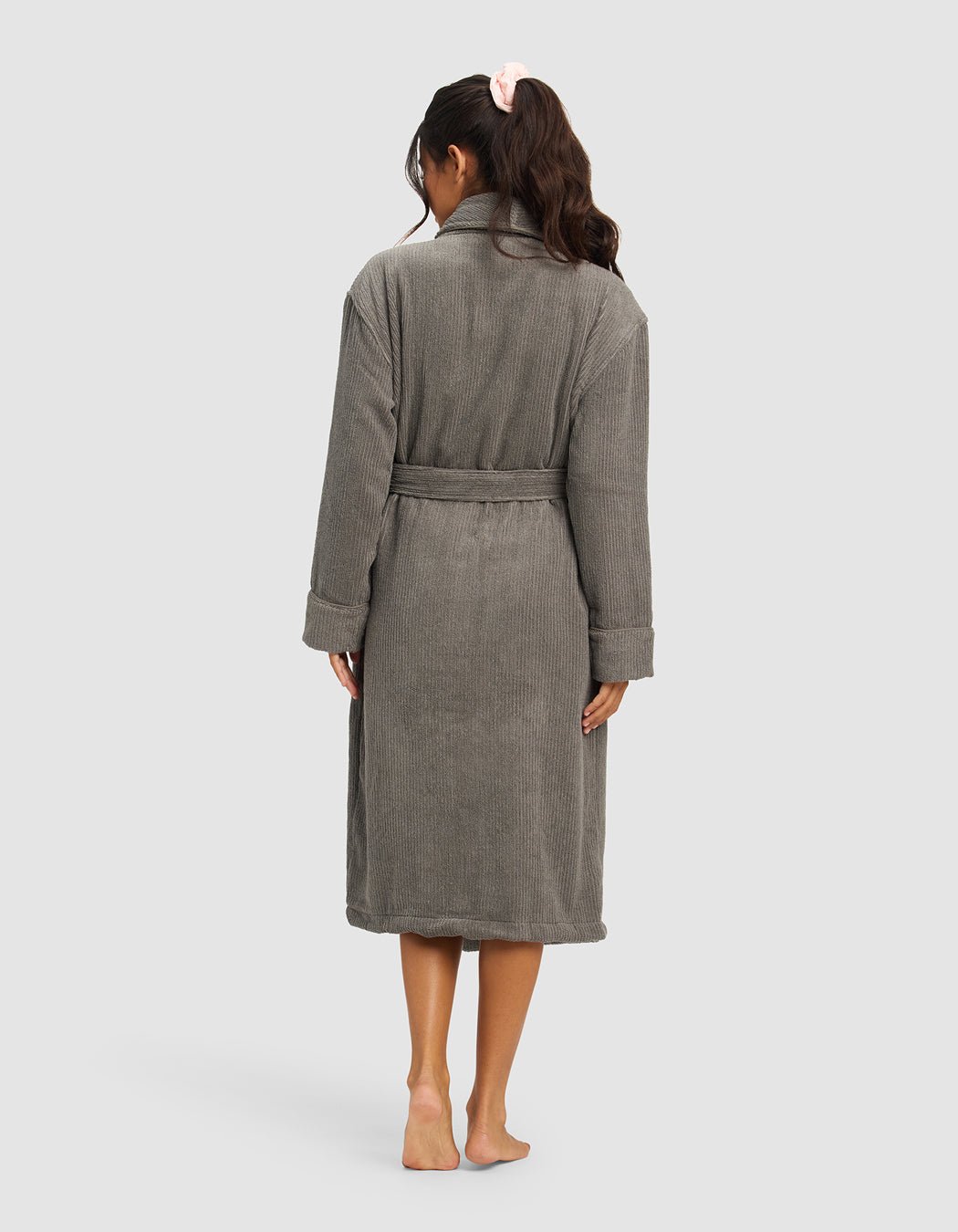 A woman stands barefoot facing away, wearing Cozy Earth's gray Ribbed Terry Bath Robe with a belt. Her long, wavy hair is tied back with a pink scrunchie against a plain light gray background.