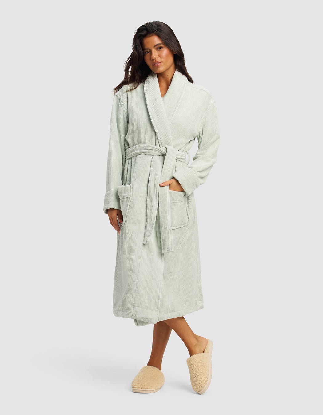 A woman with long dark hair stands against a plain background wearing the Cozy Earth Ribbed Terry Bath Robe in light sage green, featuring a belted waist and large pockets, paired with beige fuzzy slippers. She looks relaxed and comfortable.