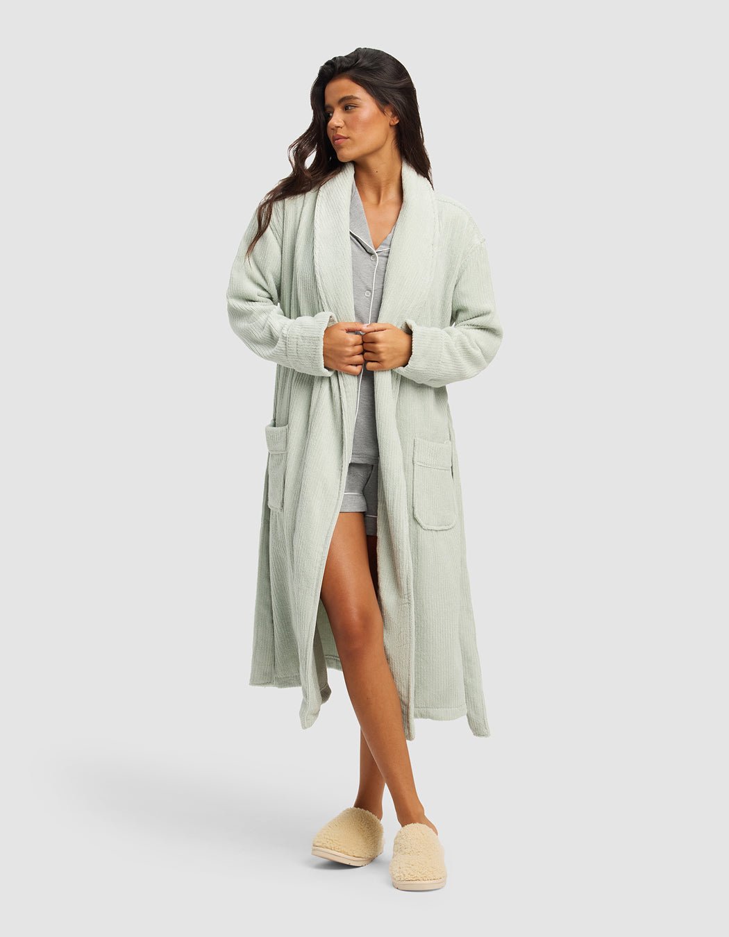A woman wears the Cozy Earth Ribbed Terry Bath Robe in light green over gray pajamas, paired with beige slippers. She stands with her hands in front, looking to the side against a plain light gray background.|Color:Laurel