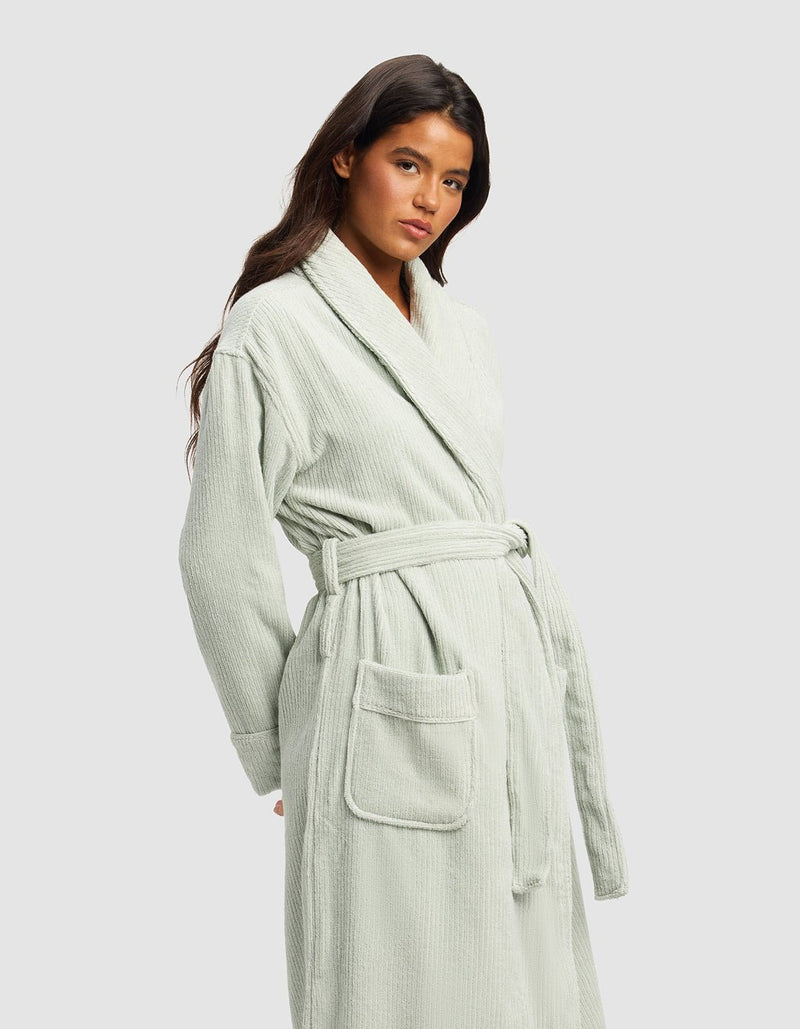 A woman with long dark hair wears the Cozy Earth Ribbed Terry Bath Robe in light sage green, featuring a belted waist and large front pockets, standing against a plain light gray background.