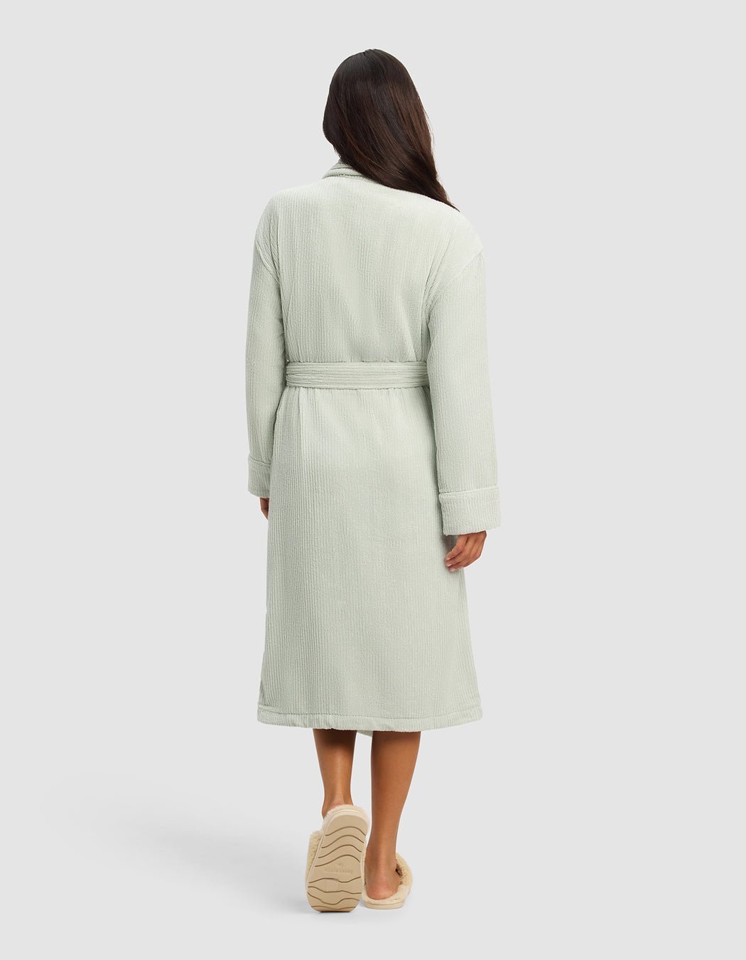 A woman faces away, wearing the Cozy Earth Ribbed Terry Bath Robe in light green with a belt and slippers, set against a plain, light-colored background.