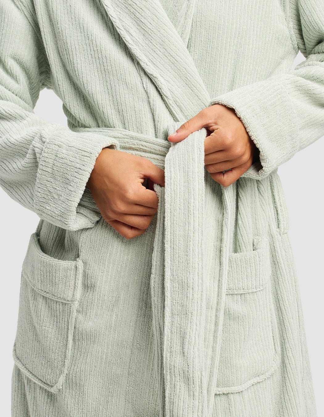 A person ties the belt of a Cozy Earth Ribbed Terry Bath Robe in light green. The textured robe features large front pockets and visible ribbing, with their hands adjusting the waist for comfort.
