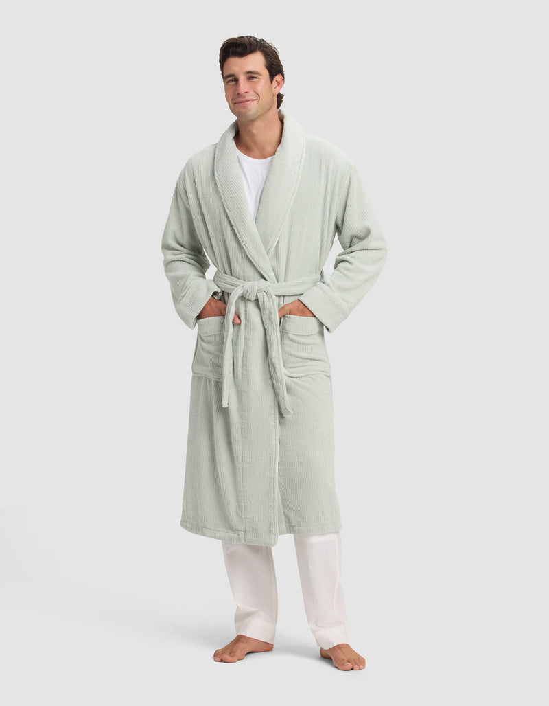 A man smiles barefoot in a Cozy Earth Ribbed Terry Bath Robe—light green with a belt and two pockets—styled over a white shirt and pants, set against a plain light background.
