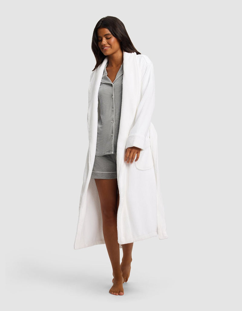 A woman with long dark hair, barefoot and smiling, models the Cozy Earth Ribbed Terry Bath Robe over gray pajama shorts and a button-up top, walking against a plain light gray background.