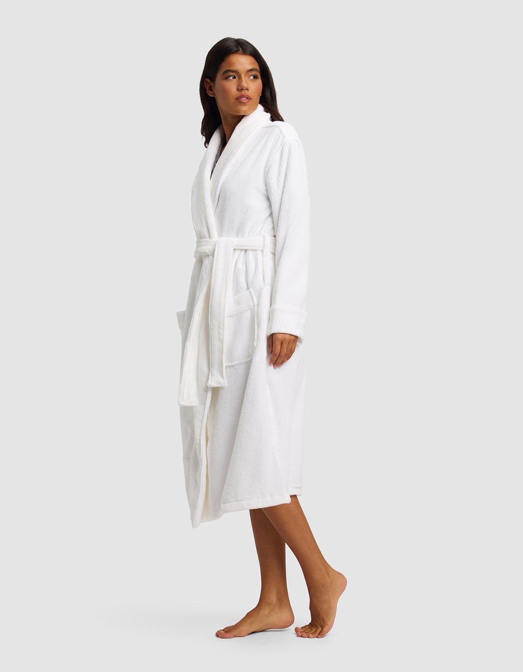 A woman stands barefoot, wearing the Cozy Earth Ribbed Terry Bath Robe—a white, knee-length robe with a belt and large front pockets—looking slightly left against a plain light gray background.