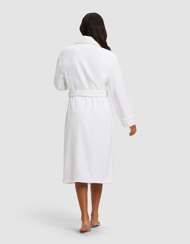 A person with long dark hair stands barefoot, facing away, wearing Cozy Earth's white Ribbed Terry Bath Robe, belted at the waist, against a plain light gray background.