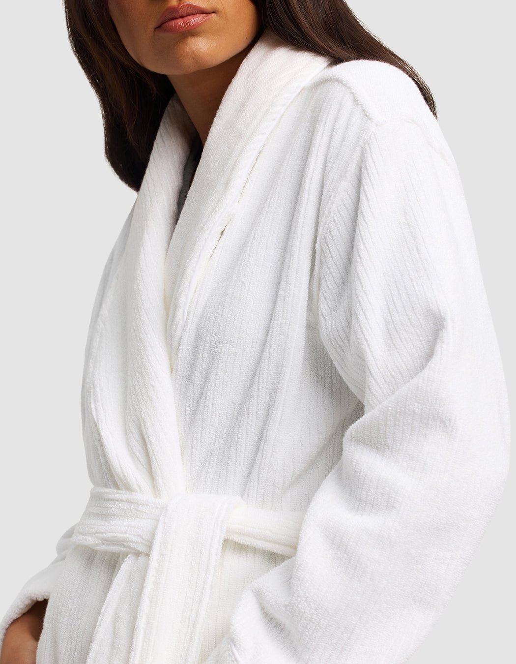 A woman models the Cozy Earth Ribbed Terry Bath Robe, featuring a plush white fabric, shawl collar, and belt. She stands against a plain light background with only her lower face and upper body visible.