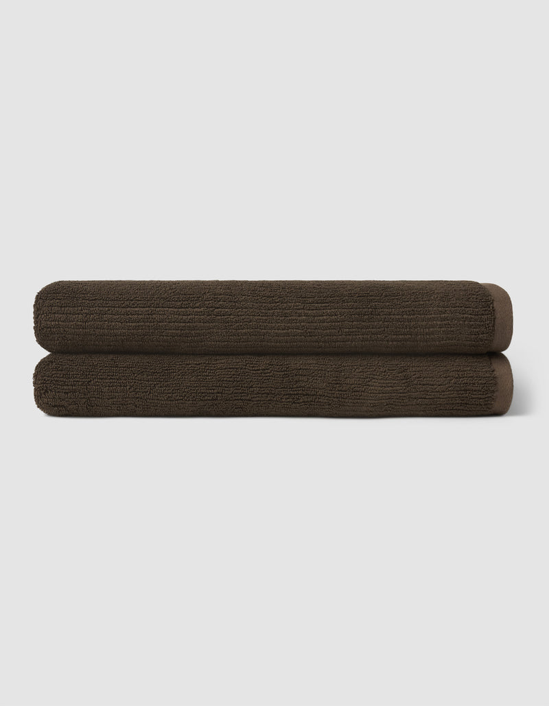 Two dark brown towels from the Cozy Earth Complete Ribbed Terry Bath Bundle are neatly rolled and stacked atop each other against a plain light gray background.