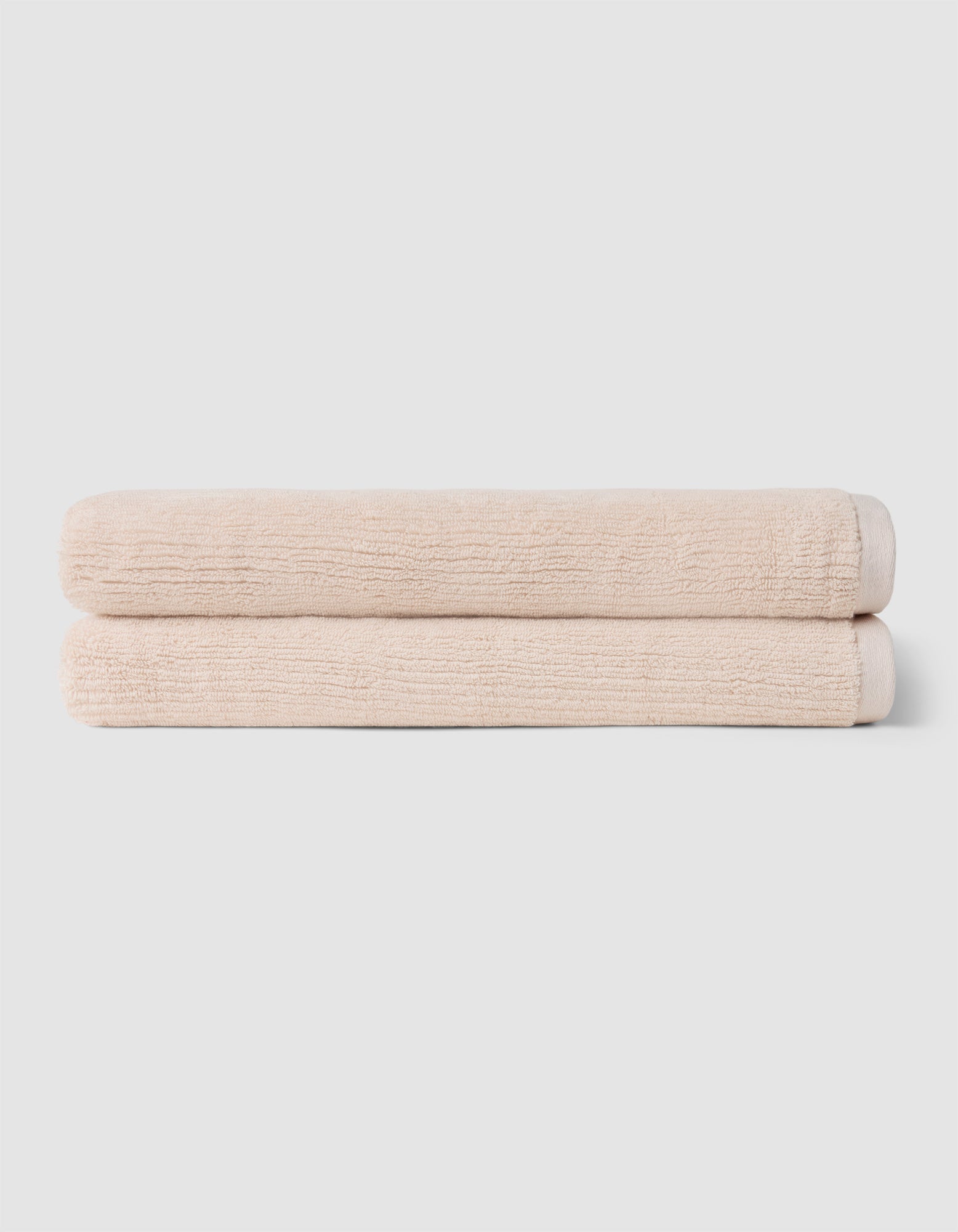Two neatly folded beige towels from Cozy Earth's Complete Ribbed Terry Bath Bundle are stacked on top of each other against a plain light gray background.|Color:Petal