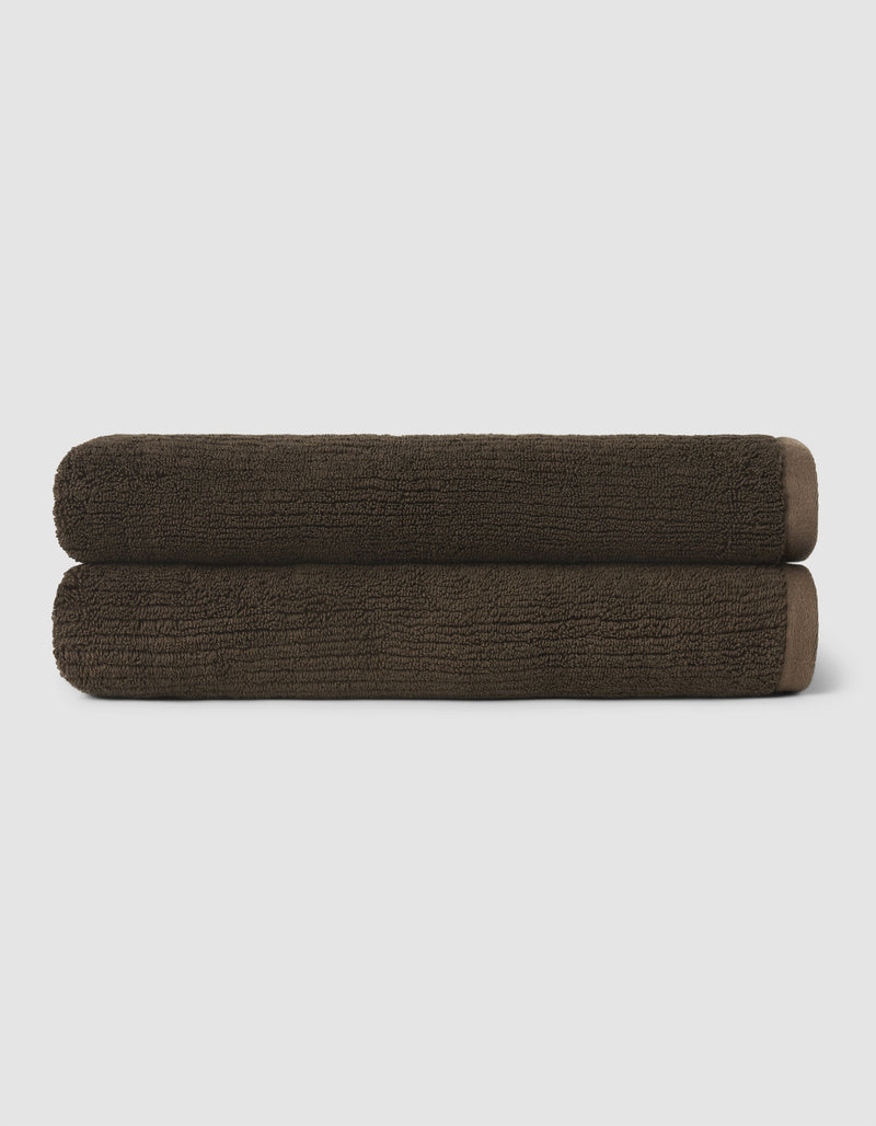 Two neatly rolled dark brown towels from the Cozy Earth Complete Ribbed Terry Bath Bundle are stacked on top of each other against a plain, light background.