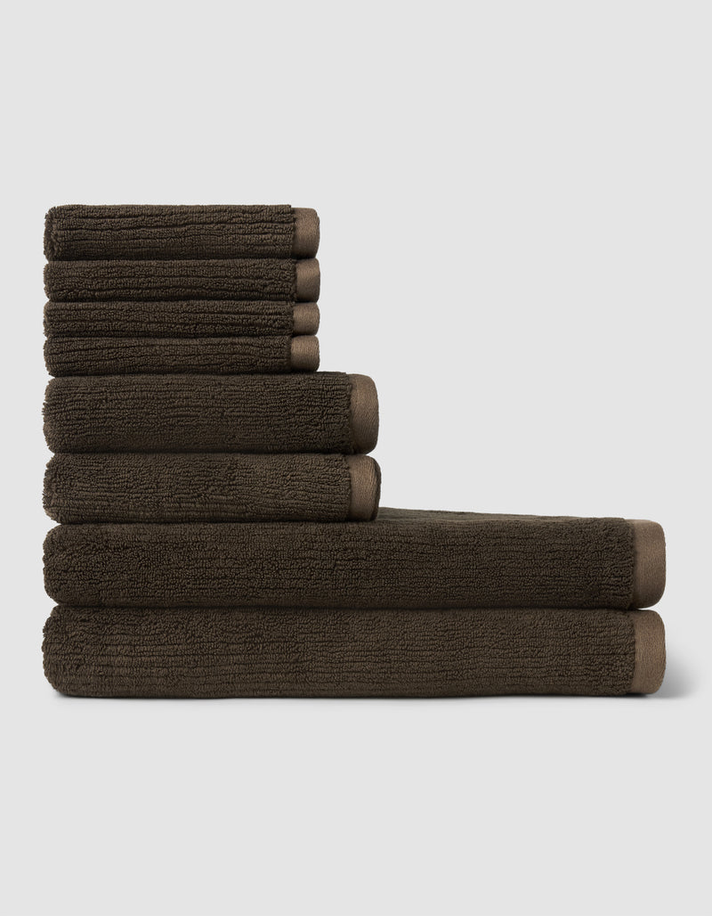 The Cozy Earth Ribbed Terry Bath Towel Set, in dark brown, features neatly stacked towels of various sizes arranged in two rows against a plain light gray background.
