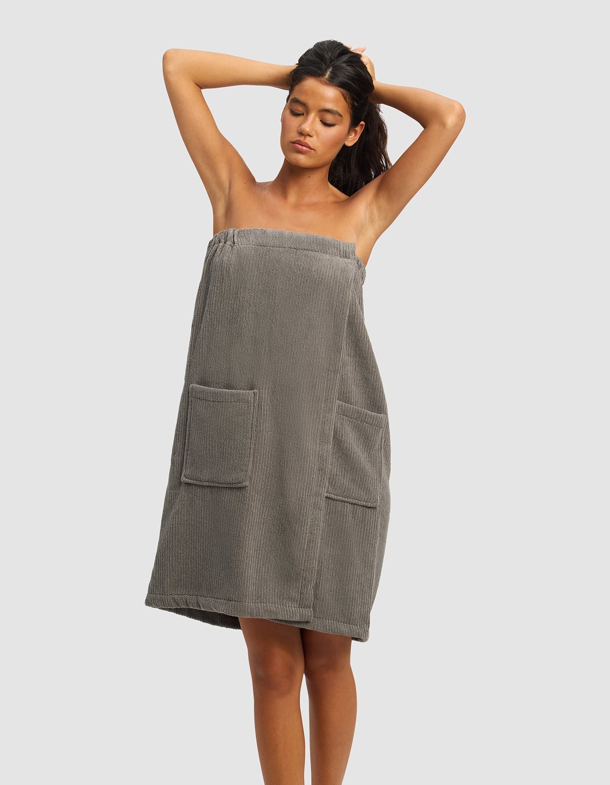 A woman stands with her hands behind her head, wearing the gray Ribbed Terry Bath Wrap by Cozy Earth, featuring a strapless design and two front pockets. The background is plain and light-colored. 