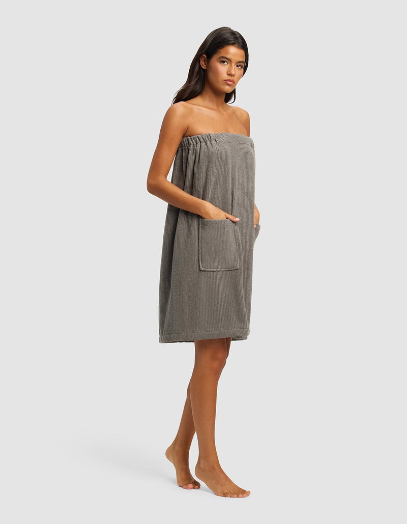 A woman stands barefoot against a plain background, wearing the Cozy Earth Ribbed Terry Bath Wrap in grey. Its strapless design features pockets, and she rests her hands in them. Her straight dark hair frames her neutral expression.