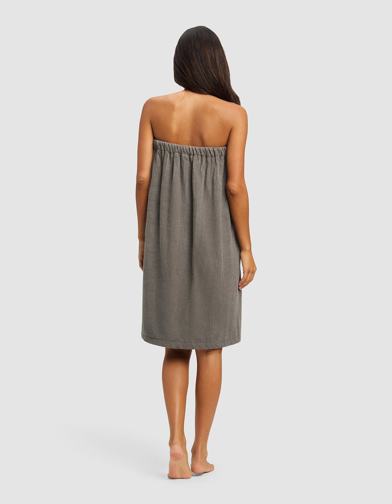 A woman with long brown hair stands barefoot, facing away from the camera, wearing the Cozy Earth Ribbed Terry Bath Wrap—a gray, strapless, knee-length towel dress—against a plain light background.