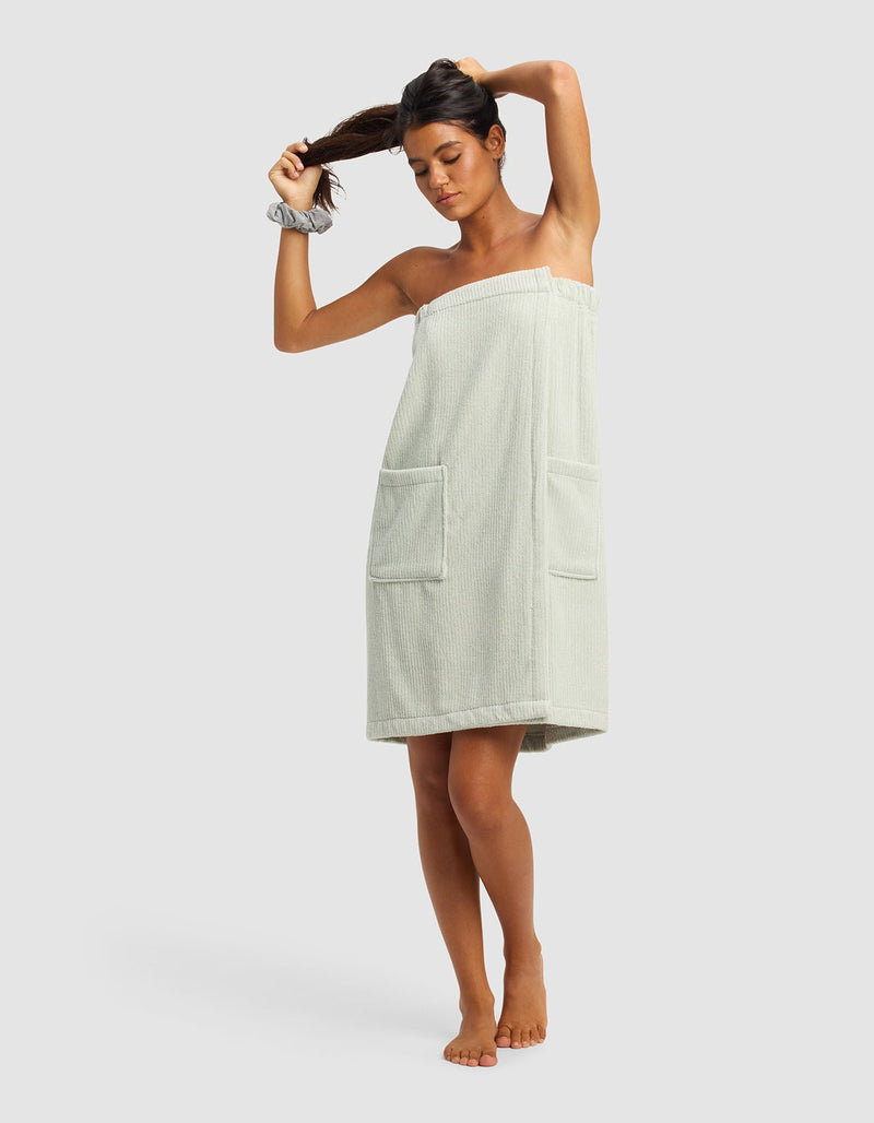 A woman stands barefoot against a plain background, adjusting her ponytail while wearing the Cozy Earth Ribbed Terry Bath Wrap in light green with two front pockets and a matching scrunchie on her wrist.