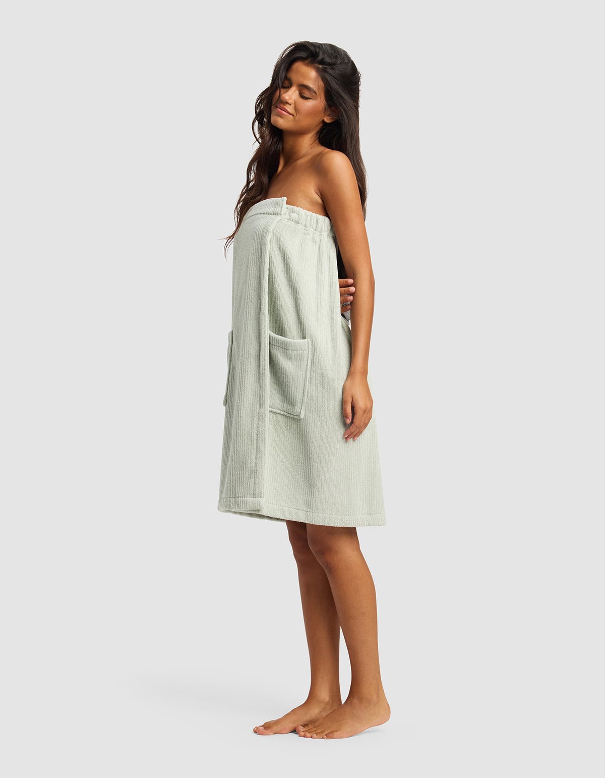 A woman with long dark hair stands barefoot, modeling the Cozy Earth Ribbed Terry Bath Wrap—light-colored, strapless, with two large front pockets—against a plain light gray background.
