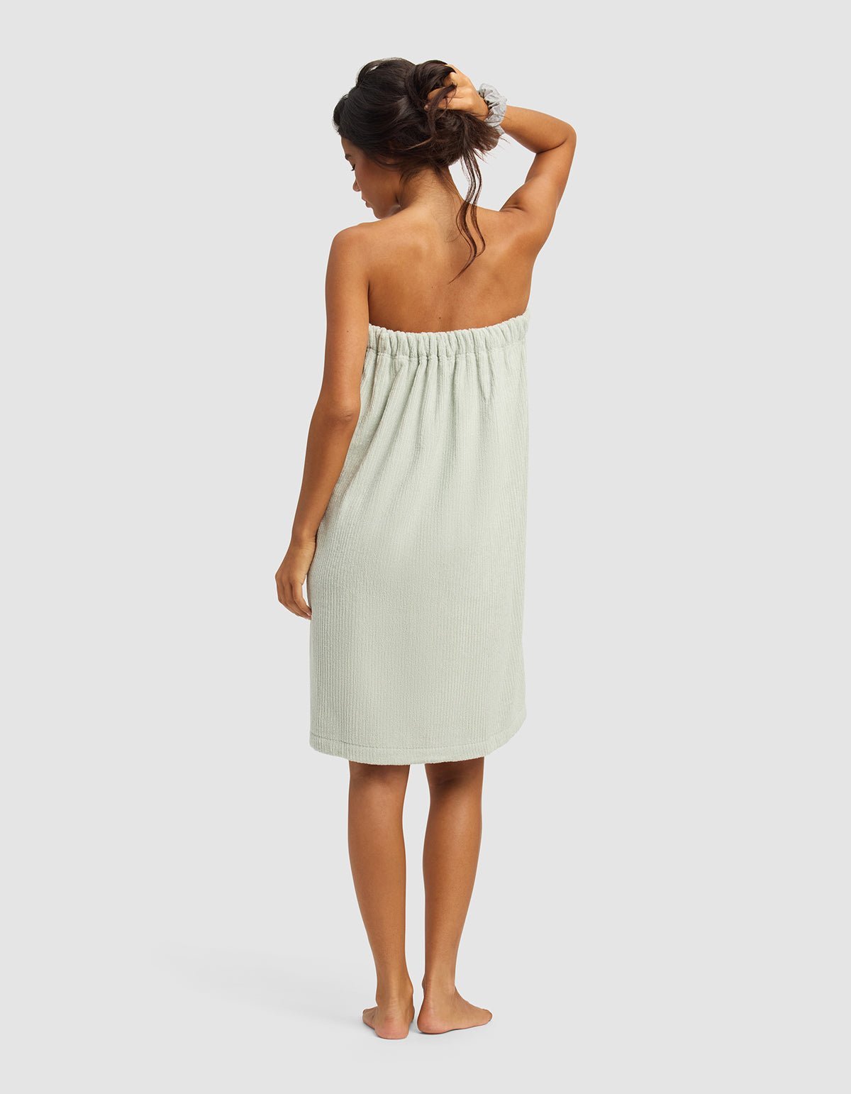 A woman stands barefoot with her back to the camera, wearing the Cozy Earth Ribbed Terry Bath Wrap in light green and a hair scrunchie, one arm raised to touch her hair, set against a plain light background.|Color:Laurel