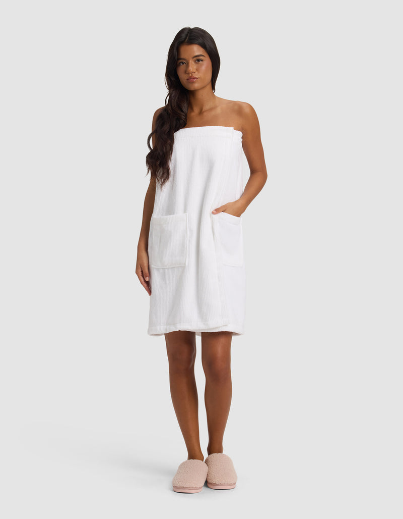 A woman with long dark hair stands facing forward, wearing the Cozy Earth Ribbed Terry Bath Wrap in white, featuring two large front pockets. She has a neutral expression and is set against a plain light gray background.