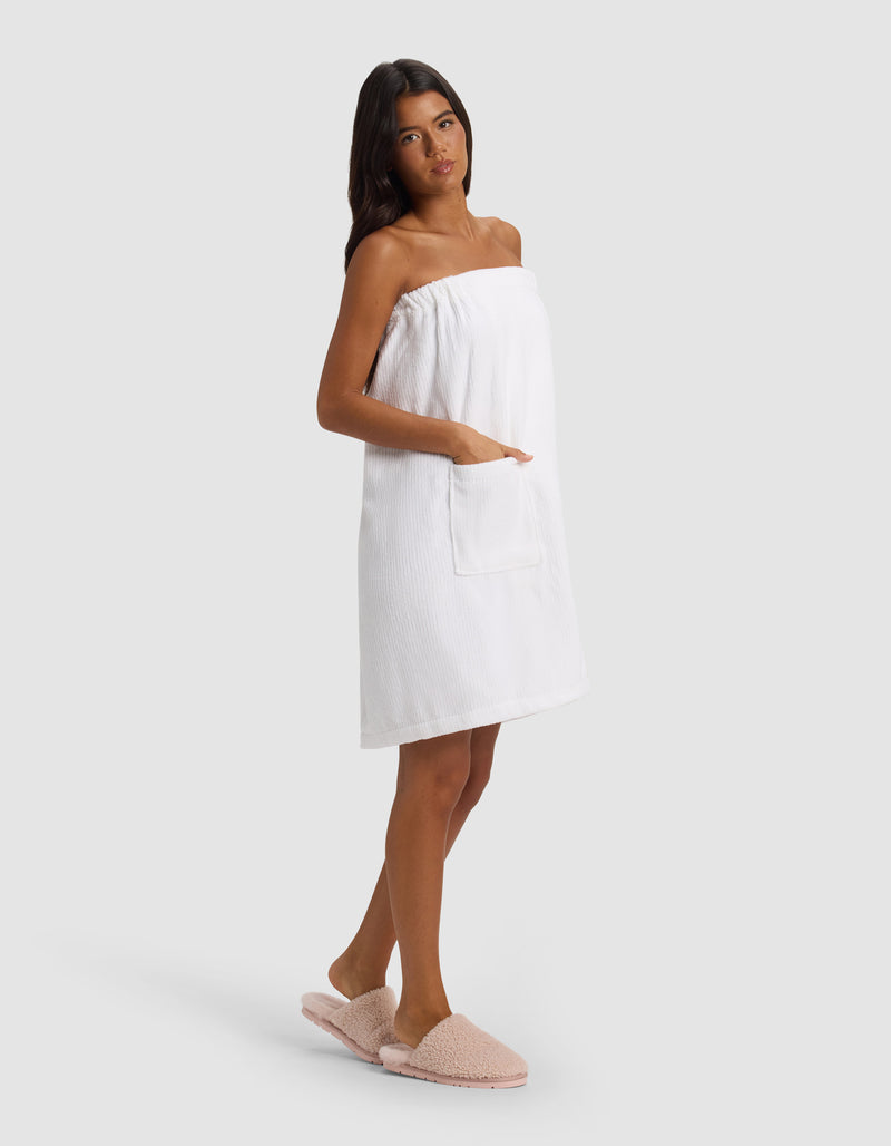 A woman with long dark hair models the Cozy Earth Ribbed Terry Bath Wrap, featuring a front pocket, paired with light pink slippers. She stands slightly turned against a plain light gray background.