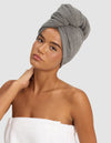 A woman poses against a plain light background, wearing the Cozy Earth Ribbed Terry Hair Towel on her head and a white towel around her body, with one hand gently touching her neck.|Color:Charcoal