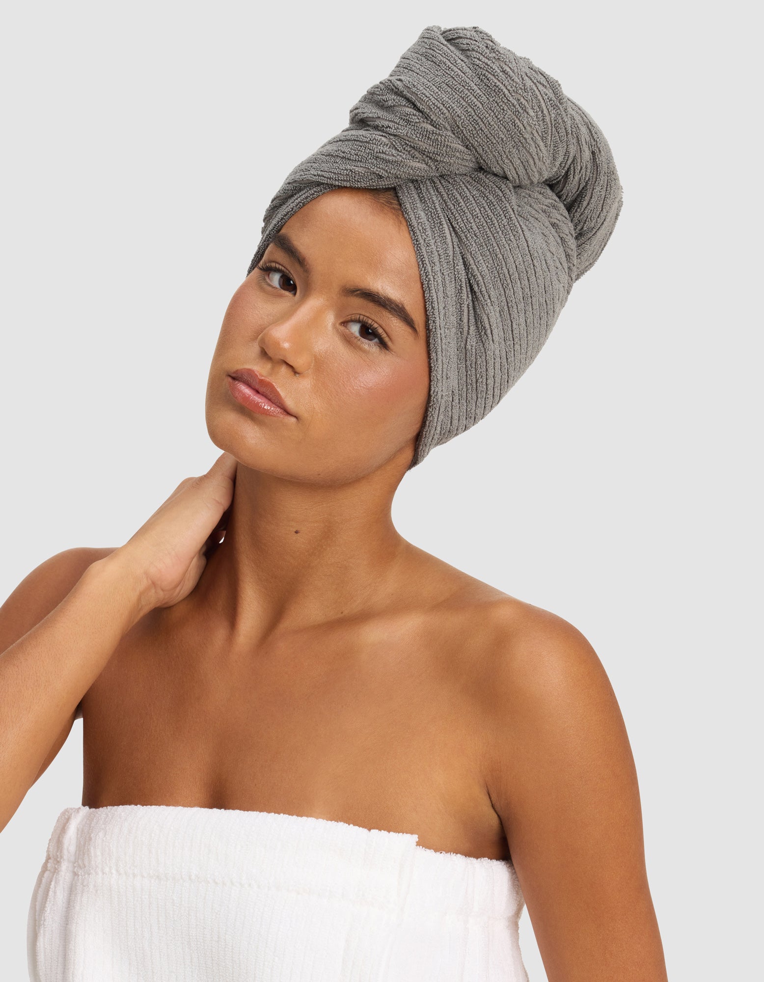 A woman poses against a plain light background, wearing the Cozy Earth Ribbed Terry Hair Towel on her head and a white towel around her body, with one hand gently touching her neck.|Color:Charcoal