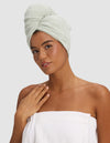 A woman with a Cozy Earth Ribbed Terry Hair Towel in light green wrapped around her head and a white towel around her chest poses against a plain background, gently touching her collarbone and smiling softly.|Color:Laurel