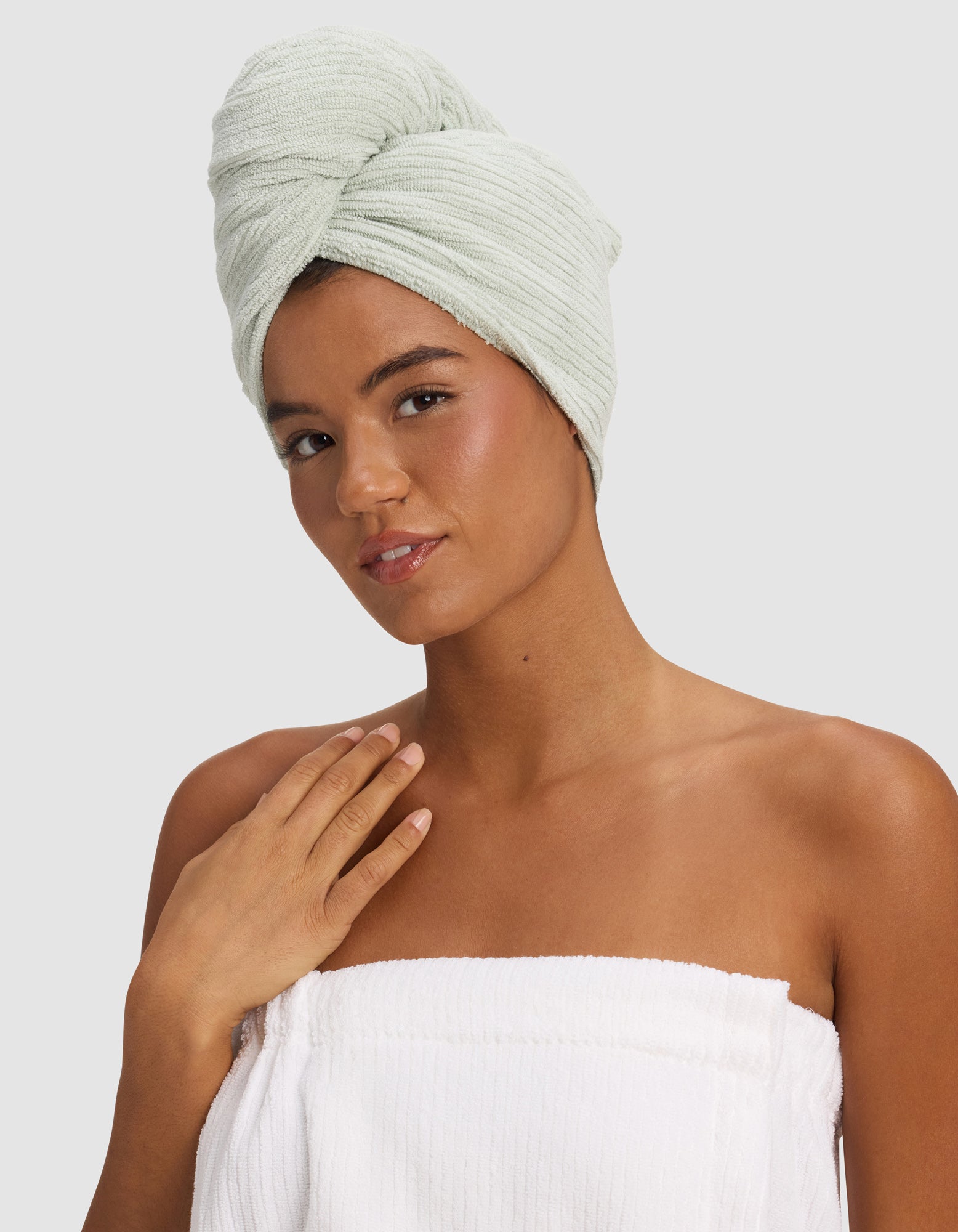 A woman with a Cozy Earth Ribbed Terry Hair Towel in light green wrapped around her head and a white towel around her chest poses against a plain background, gently touching her collarbone and smiling softly.|Color:Laurel