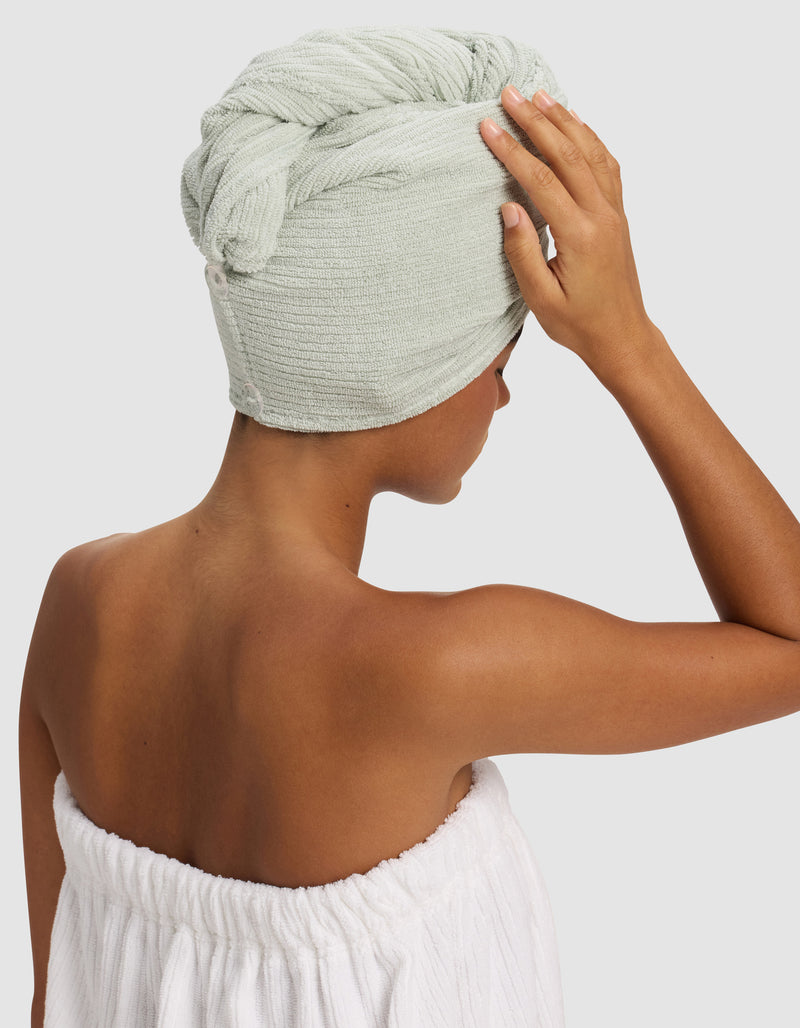 A person with medium brown skin wears a Cozy Earth Ribbed Terry Hair Towel in light green wrapped around their head and a white towel around their body, facing away with one hand touching their head.