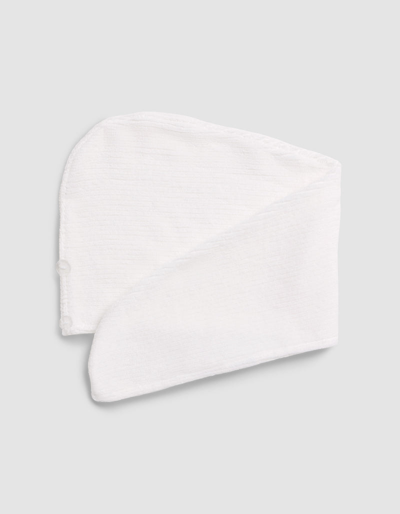 The Cozy Earth Ribbed Terry Hair Towel, in white textured fabric, is folded flat on a light gray surface. It features a curved top, long tapered end, and a small side button.